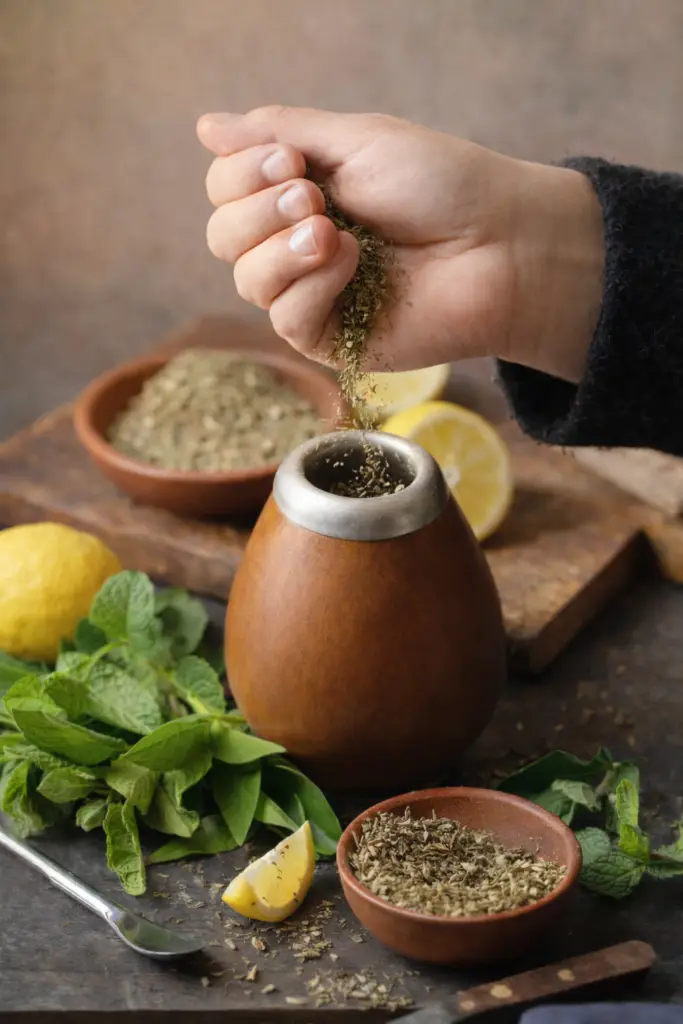 Mão adicionando ervas secas em recipiente tradicional com plantas frescas ao redor