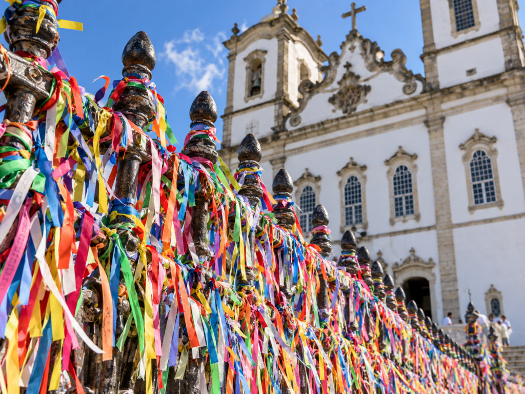 Muitas fitas coloridas amarradas a um gradil de ferro, diante de uma igreja de arquitetura barroca em uma cena ensolarada.